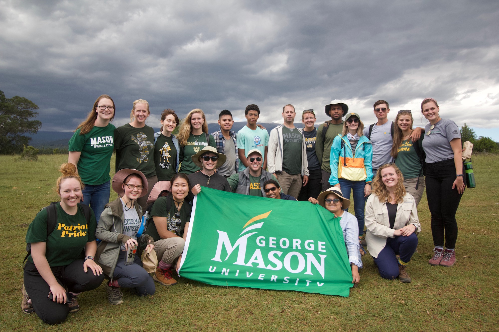 Graduate and undergraduate students in Mason attire holding a green flag that says George Mason University